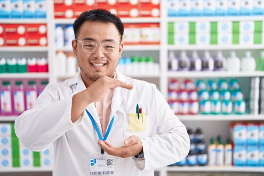 Chinese young man working at pharmacy drugstore gesturing with hands showing big and large size sign, measure symbol. smiling looking at the camera. measuring concept.