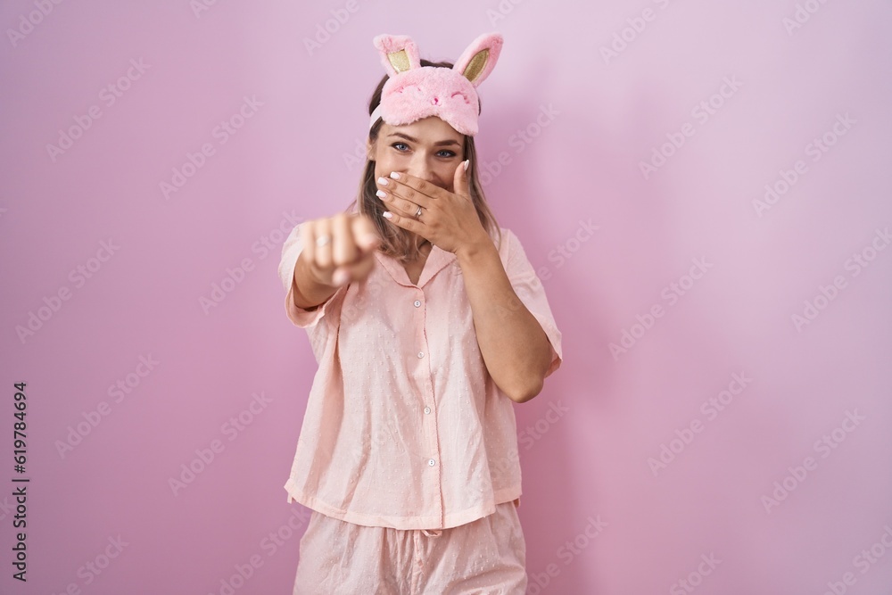 Sticker blonde caucasian woman wearing sleep mask and pajama laughing at you, pointing finger to the camera 