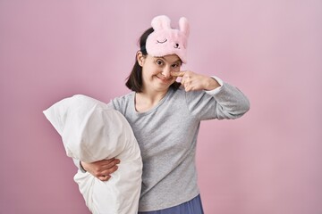 Woman with down syndrome wearing sleeping mask hugging pillow pointing with hand finger to face and nose, smiling cheerful. beauty concept