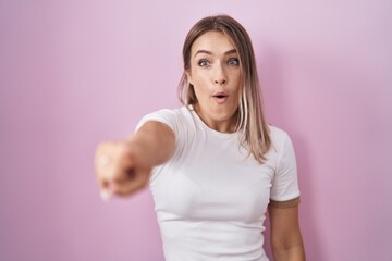 Blonde caucasian woman standing over pink background pointing with finger surprised ahead, open mouth amazed expression, something on the front