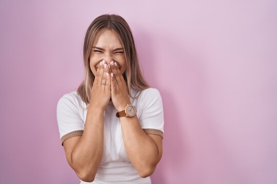Blonde caucasian woman standing over pink background laughing and embarrassed giggle covering mouth with hands, gossip and scandal concept