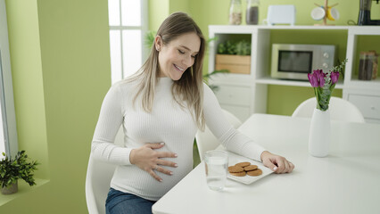 Young pregnant woman eating cookies sitting on table at dinning room