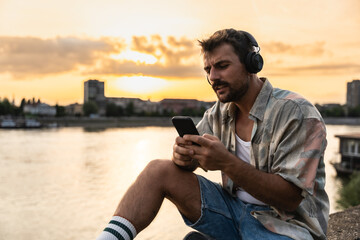 Young hipster happy man sitting near the river in the city escaping from city life listening music...