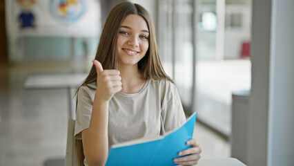 Young beautiful girl student reading book doing thumb up gesture smiling at library