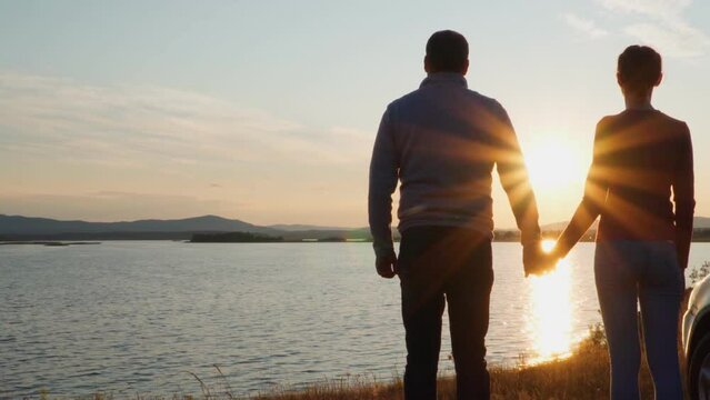 A man and a woman stand on the shore of the lake near their car and admire the sunset, holding hands. The golden hour.