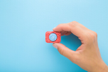 Young adult woman hand holding and showing blue red dishwasher tablet on light blue table background. Pastel color. Closeup. Top down view.