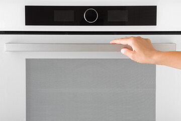 Young adult woman hand holding handle and opening glass door of modern light gray oven with black touch screen display. Front view. Closeup.