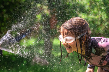 Happy girl splash and plays with garden hose with sprinkler in backyard.