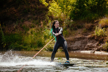 A girl in a wetsuit stands on a wakeboard on the river