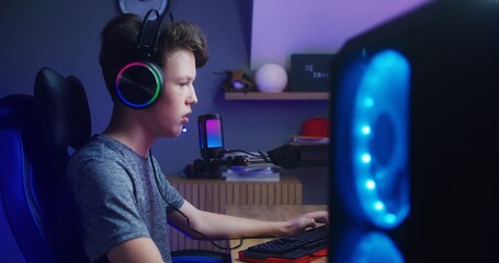 Concentrated boy in headphones plays in multiplayer game on PC at home. Gamer talks with teammates using microphone. Online video game live streaming or esports tournament. Side view.