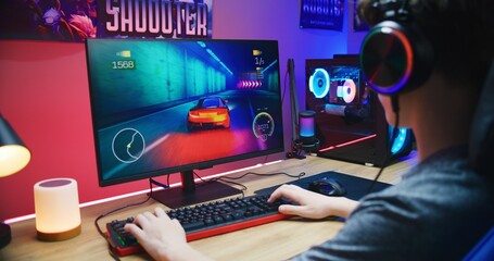 Young gamer plays in car racing simulator on PC at home. Computer monitor with displayed online video game live stream or cybersport championship. Desk illuminated by RGB LED strip light.