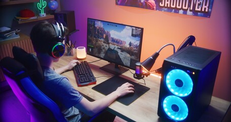 Young gamer controls tank while playing game on PC at home. Computer monitor with displayed online video game live stream or cybersport tournament. Desk illuminated by RGB LED strip light. Back view. © Framestock