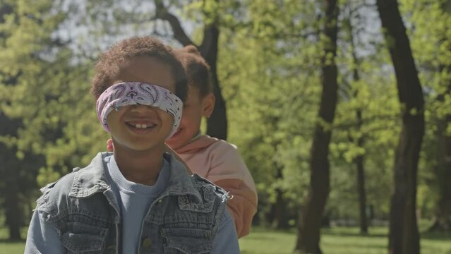 Little African American girl blindfolding smiling friend while preparing to play blind mans buff together in park on summer day