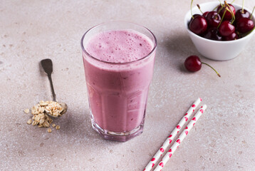 Milkshake with cherries, yogurt and oatmeal, drinking straws, bowl with fresh cherries on a beige textured background