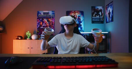 Young gamer in VR headset plays virtual online video game using wireless controllers. Boy enjoys eSports tournament. Room with warm neon LED lights. Gaming at home. POV from PC screen perspective. - obrazy, fototapety, plakaty