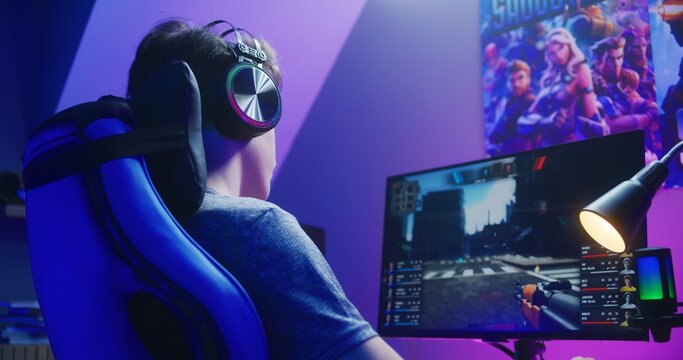 Young gamer in headphones plays in first person shooter on computer and loses round in PvP multiplayer game. Online video game live streaming or esports competition. Gaming at home concept. Back view.