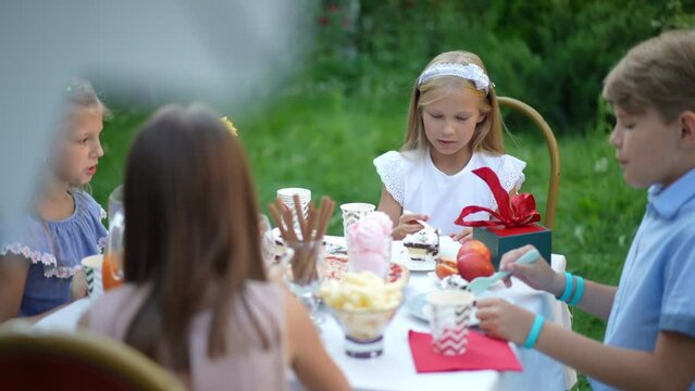 Blond girl in white dress eating sweet cake sitting with friends on backyard celebrating birthday. Portrait of relaxed happy Caucasian child enjoying leisure and tasty food outdoors