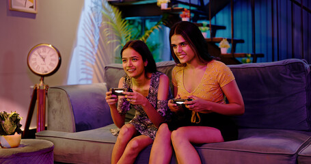 Happy Asian Indian two teen girl adult women couple playing video game hold joypad in hand sitting sofa couch making funny playful moment enjoy fun joy weekend late night at indoor interior home house