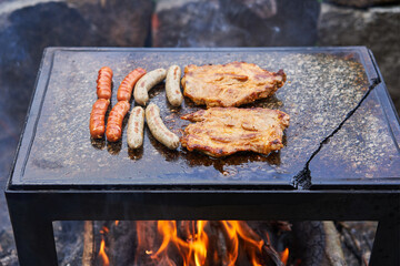Traditional old fashion barbeque. Bavarian style and normal sausages with two pork collar steaks on grill made from granit stone on the open fireplace in the summer evening. Delicious, and tasty food.