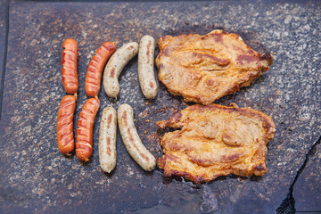 Detailed top view on the granite stone grill with two kind of sausages and pork collar steaks. On the open fireplace in the summer evening. Delicious, and tasty food perfect for party with friends.
