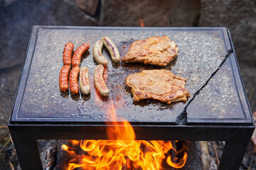 Old fashion style summer barbeque tasty party food. Two kinds of sausages, spicy sausages and white bavarian ones and two steaks from marinated pork collar on the granit stone slab over the camp fire.