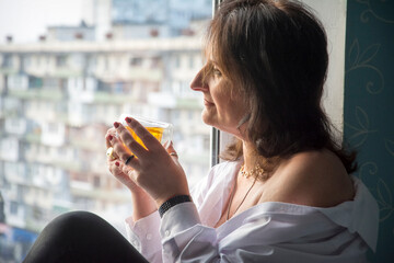 A woman with a cup of tea sits on a window sill during the day.