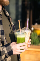 Fototapeta premium A man outdoors holds plastic cups with green and orange smoothies, a delicious cooling summer drink.