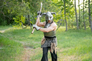 Tuinposter Portrait of a warrior in a horned helmet, a steel breastplate with a two-handed ax in his hands, posing against the backdrop of the forest.   © Михаил Гута