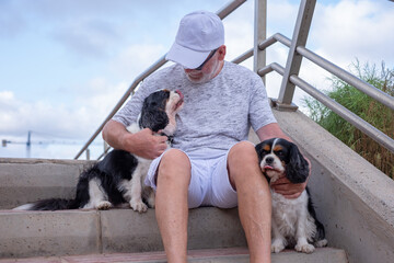 Portrait of senior caucasian man in white cap sitting on outdoors staircases with his two cavalier king charles dogs. Best friend forever concept