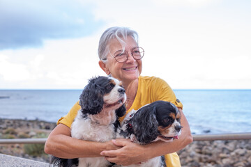 Smiling senior woman in yellow jersey sitting close to the beach embracing her two cavalier king...