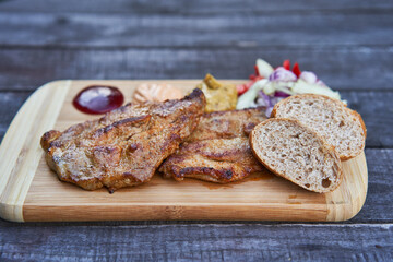 Two grilled barbeque pork collar steaks served outside in garden restaurant on the wooden plate with slices of whole grain bread, choped onions, tomatoes, mustard and barbeque sauces. Tasty party food