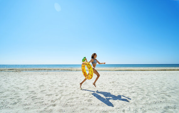 Paradise seascape of stunning panoramic image.Bronzed joyful young woman running along the endless sandy beach, carrying a pineapple float ring, ready to dive into the azure water.