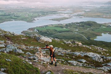 Young fit atheltic man with backpack stand on mountain top during sunset or midnight sun in Norway. Summer evening hikes. Put stone on top of cairn. Hiking in scandinavia and nordic countries
