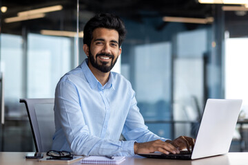 Portrait of happy and successful businessman, indian man smiling and looking at camera, satisfied with achievement results man working inside office building using laptop at work