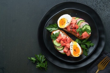 Healthy sandwich with bread or toast, smoked salmon, soft egg, cream cheese, cucumber, radish, black sesame and parsley on black plate. Delicious protein fish sandwich for breakfast. Smorrebrod.