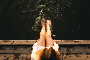 Woman relaxes by the lake sitting on the edge of a wooden jetty, swing feet near the water surface,...