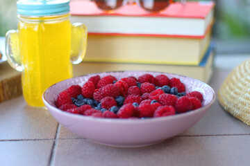 Straw hat, orange soda in the cactus shaped cup, bowl of fresh blueberreis and raspberries, plate of chocolate chip cookies, stack of books and sunglasses in the garden. Selective focus.