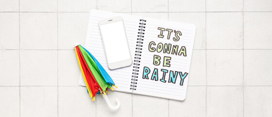 Notebook with text IT'S GONNA BE RAINY, mobile phone with blank screen and small umbrella on white tile background