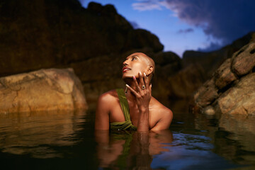 Non-binary person of color poses in water, shows jewelry inside scenic creek at night. Lgbtq ethnic...