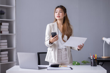 Corporate communication. Happy asian female manager working with papers and using phone, reading financial documents while sitting at workplace in office.