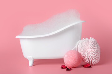 Small bathtub with foam, sponge and bath bomb on pink background