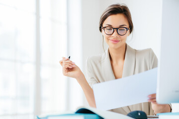 Concentrated businesswoman studies contract terms, writes with pen, dressed formally, at desk in spacious white interior