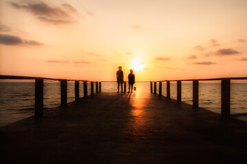 Silhouette of two people standing with their backs turned and talking. At the end of the beach bridge during sunset time.