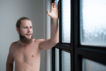 Handsome young man standing near the window and looking out the window