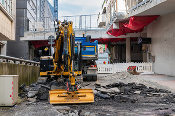 Yellow excavator and truck on a construction site between buildings. Narrow city street.