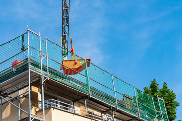 Lifting crane holds an orange container over the roof of a renovated house. Scaffolding green mesh...