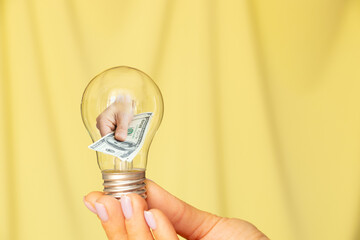 A woman's hand holds a light bulb and the second hand gives dollars from a light bulb on a yellow background, income and profit