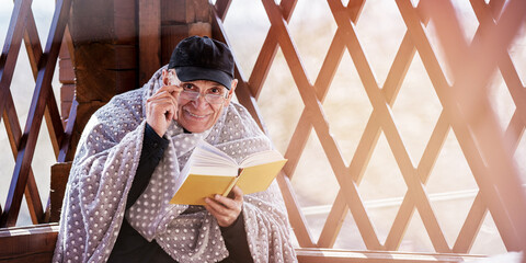 Grandfather wrapped in warm blanket reads book while sitting in wooden gazebo in countryside