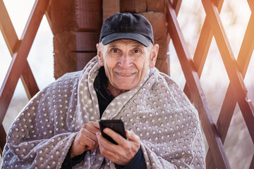 Senior person wearing cap and warm plaid with smartphone in hands sitting in wooden gazebo in cottage