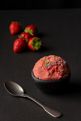 Top view of strawberry ice cream in bowl and black table with spoon and strawberries, vertical, with copy space
